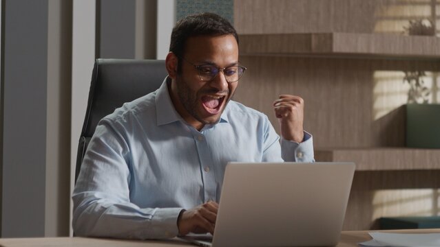 Happy cheerful Indian business man celebrating victory at office laptop winning American executive businessman lucky male excited good news success online bet win achievement computer triumph indoors