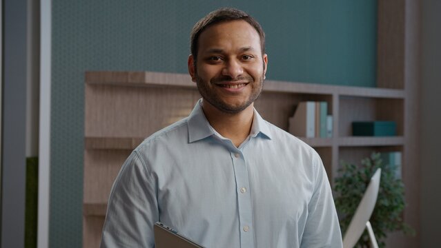 Joyful Indian Arabian business man smiling looking camera indoor office laptop workplace happy American businessman entrepreneur employer manager holding computer corporate portrait marketing company