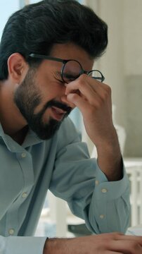 Exhausted Arabian Indian business man rubbing dry eyes working laptop indoor office tired male businessman worker guy headache bad vision removing glasses overwork desk computer burnout health problem