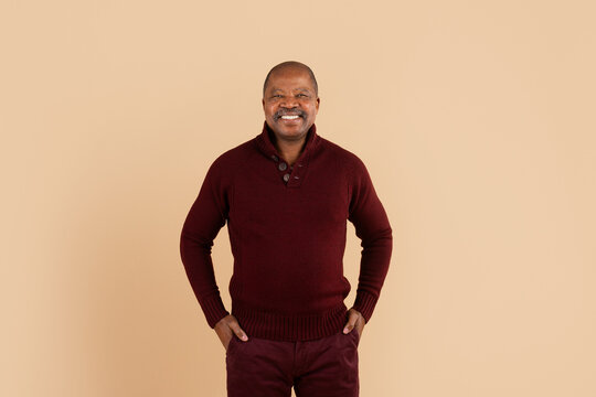Smiling mature African American man standing confidently with hands in pockets on beige studio background, wearing burgundy sweater in relaxed lifestyle portrait with friendly expression