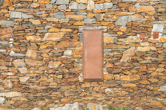 Rustic Stone Wall with Rusty Metal Door, Wooden Lintel, and Ventilation Grilles in Warm Earth Tones