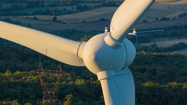 Aerial view of a wind turbine illuminated by warm morning light, with a high-speed train, power lines and agricultural landscape in the background in Marsanne, Auvergne-Rhone-Alpes, France.