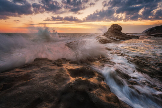 View of the Sedia del Papa rock formation with waves crashing against the rocky coastline during a dramatic sunset in Ancona, Marche, Italy.