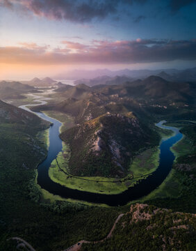 Aerial view of the winding Rijeka Crnojevica river bend surrounded by lush green hills and mountains under a soft sunset sky in Rijeka Crnojevica, Cetinje Municipality, Montenegro.