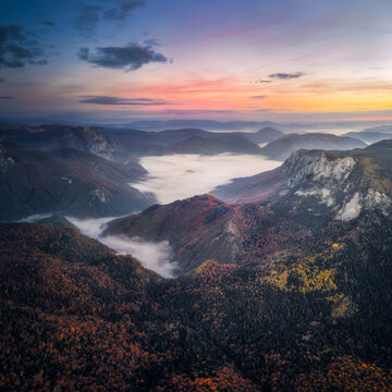 Aerial view of Sutjeska National Park with autumn forests, rugged mountain peaks, and fog in the valley under a colorful sunset sky in Tjentiste, Republika Srpska, Bosnia and Herzegovina.