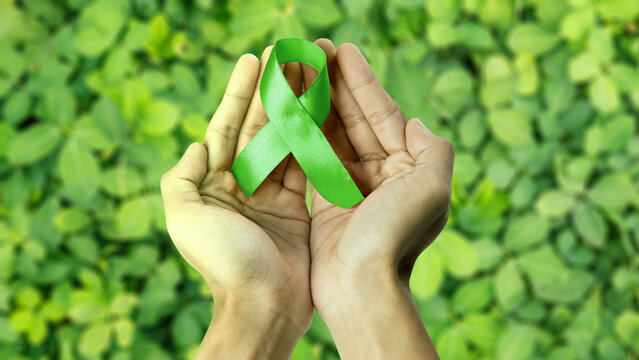 Hands holding a green awareness ribbon, symbolizing mental health or environmental support, against blurred green foliage. Concept of World Mental Health Day