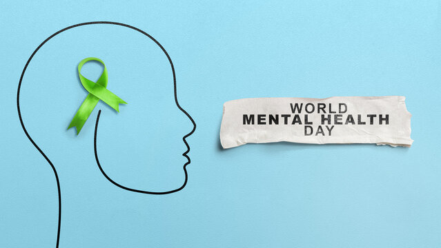 A green awareness ribbon inside a black drawn human head outline next to a torn paper with World Mental Health Day text on a blue background. Concept of World Mental Health Day