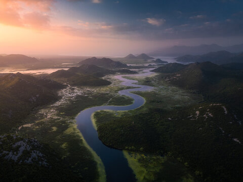 Aerial view of the winding Crnojevica River flowing through lush green mountains and wetlands during golden hour in Rijeka Crnojevica, Cetinje Municipality, Montenegro.