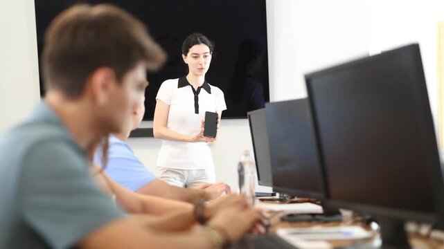 Enthusiastic young girl holding smartphone and pointing to screen while presenting educational mobile app to group of fellow students in computer lab. High quality 4k footage