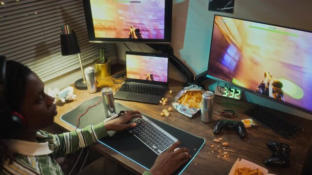 African American teenager playing shooter video game on multiple screens at messy desk. POV high angle shot suitable for esports marketing or gaming addiction awareness campaigns