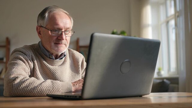An elderly man sits at a table in his home. He focuses on his laptop screen while studying. Natural light comes in through the window, creating a bright atmosphere.