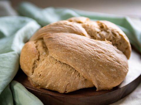Golden crusty sourdough knot bread loaf on a wooden board next to a mint green linen cloth. Close-up shot with soft blurred background and natural daylight. Great for cozy lifestyle food concepts