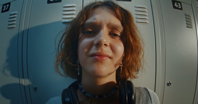 Close Up Portrait of Teenage Girl With Curly Brown Hair Wearing Black Headphones and Cross Earrings, Looking at Camera With Confused and Uncertain Expression in Front of Metal School Lockers.