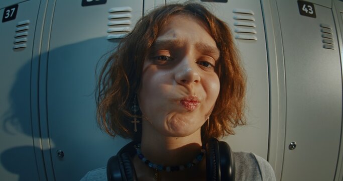 Close Up Portrait of Teenage Girl With Curly Brown Hair Wearing Black Headphones and Cross Earrings, Looking at Camera With Confused and Uncertain Expression in Front of Metal School Lockers.