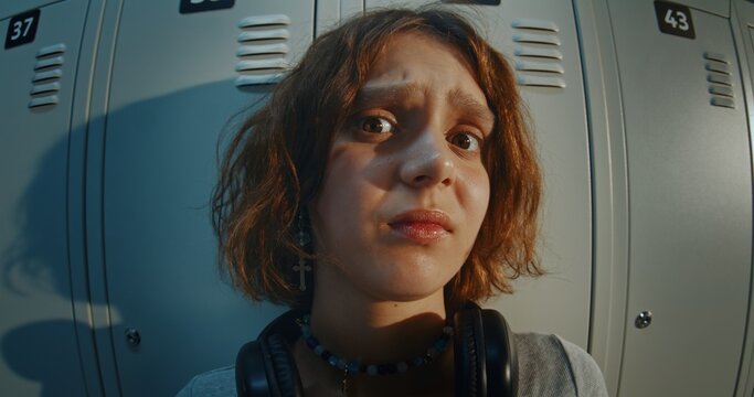Close Up Portrait of Teenage Girl With Curly Brown Hair Wearing Black Headphones and Cross Earrings, Looking at Camera With Confused and Uncertain Expression in Front of Metal School Lockers.