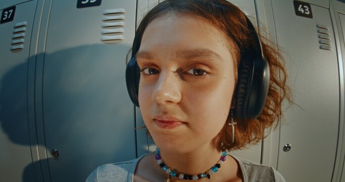 Close Up Portrait of Curly Haired Teenage Girl Wearing Headphones and Listening to Music in High School Hallway. Relaxed and Thoughtful Expression, Direct Gaze, Youth Lifestyle and Gen Z Identity.