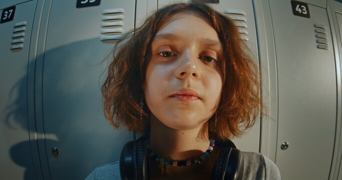 Close Up Portrait of Curly Haired Teenage Girl Wearing Headphones and Listening to Music in High School Hallway. Relaxed and Thoughtful Expression, Direct Gaze, Youth Lifestyle and Gen Z Identity.