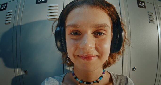 Close Up Portrait of Curly Haired Teenage Girl Wearing Headphones and Listening to Music in High School Hallway. Relaxed and Thoughtful Expression, Direct Gaze, Youth Lifestyle and Gen Z Identity.