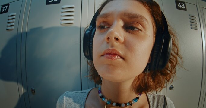 Close Up Portrait of Curly Haired Teenage Girl Wearing Headphones and Listening to Music in High School Hallway. Relaxed and Thoughtful Expression, Direct Gaze, Youth Lifestyle and Gen Z Identity.