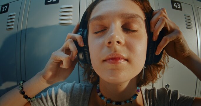 Close Up Portrait of Curly Haired Teenage Girl Wearing Headphones and Listening to Music in High School Hallway. Relaxed and Thoughtful Expression, Direct Gaze, Youth Lifestyle and Gen Z Identity.