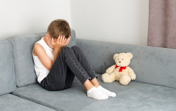 Boy crying with his head down and sitting sad scared on the sofa in his room and hugs his knees. Concept domestic violence and child abuse.