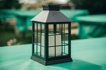 Black metal and glass lantern with a white candle stands on a turquoise-covered outdoor table, with benches and greenery softly blurred in the background and ample copy space. © Jolanta
