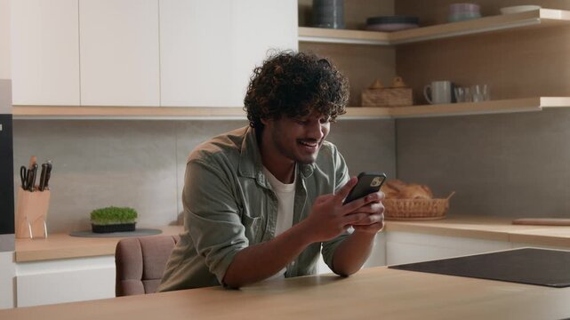 Happy Indian Arabian man celebrating smartphone victory at home kitchen winning prize reward success mobile phone excited ethnic male achievement financial cash online betting app joyful winner indoor