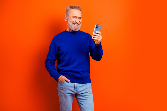 Smiling senior man in blue sweater using smartphone against bright orange background &mdash; casual portrait of happy older adult, technology, communication and modern lifestyle