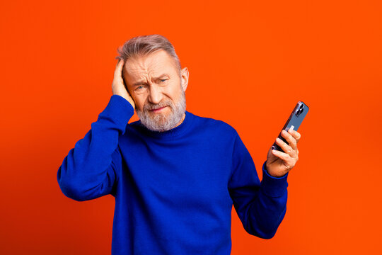 Confused senior man holding a smartphone with a frustrated expression against a bright orange background struggling with technology and communication