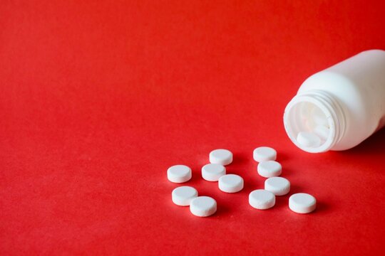 White round medical pills scattered on a solid red surface next to an open white plastic bottle.