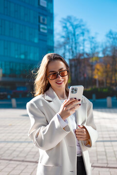 Professional businesswoman holding smartphone with confident smile outdoors. Confident woman using mobile phone near modern office building, digital communication