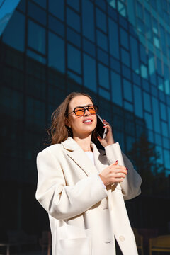 Confident businesswoman talking on smartphone with positive expression. Professional woman making phone call outside modern office building, business communication and corporate lifestyle concept.