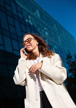 Confident businesswoman smiling during smartphone conversation outdoors. Professional woman enjoying successful phone call beside modern office building, business communication 