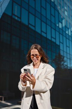 Happy businesswoman holding smartphone with smiling confident expression outdoors. Professional woman using mobile phone beside modern office building, business communication 