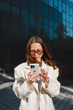 Confident businesswoman using smartphone with elegant professional style outdoors. Professional woman checks mobile phone beside modern office building, 