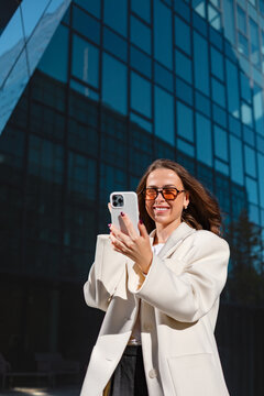 Smiling businesswoman showing smartphone with cheerful confident expression. Professional woman enjoys mobile communication beside modern office building, 