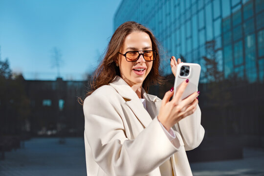 Cheerful businesswoman making video call with smartphone outdoors. Professional woman communicates beside modern office building, digital technology and successful corporate lifestyle concept.