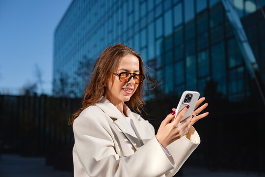 Smiling businesswoman making video call with smartphone confidently outdoors. Professional woman communicates beside modern office building, digital communication and successful corporate