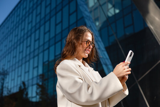 Professional businesswoman using smartphone with smiling confident expression. Confident woman checks mobile phone beside modern office building, 