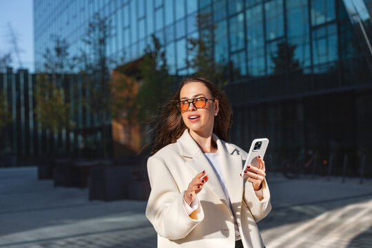 Cheerful businesswoman gesturing with smartphone during outdoor conversation. Professional woman uses mobile phone beside modern office building, business communication 