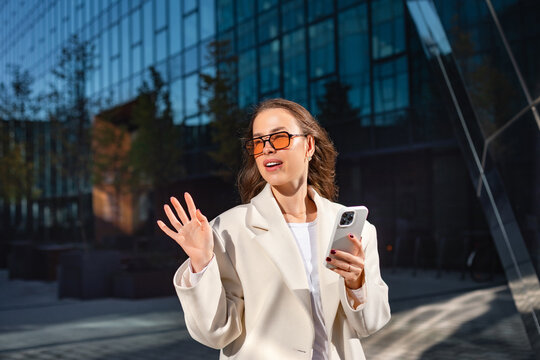 Smiling businesswoman waving while holding smartphone with confident expression. Professional woman enjoys mobile communication beside modern office building,