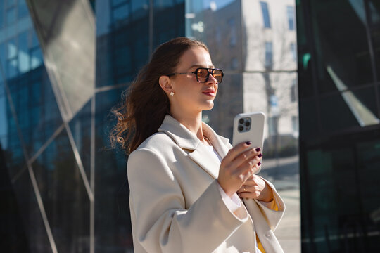 Professional businesswoman using smartphone with focused confident expression outdoors. Confident woman checks mobile phone beside modern glass office building, digital communication 