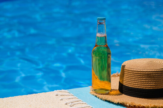 A bottle of cold beer and a straw hat by the pool.