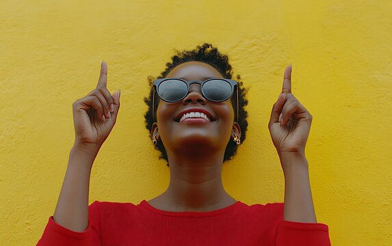 A young woman in sunglasses smiles and points upwards with both hands, against a bright yellow wall.