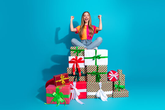 Happy young woman sits on colorful gifts and celebrates a festive season in a bold blue studio scene