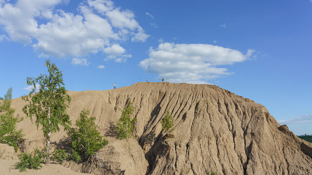 Unrecognizable people standing on a clay hilltop with distinct ridges under a blue sky with scattered clouds, surrounded by sparse vegetation and trees in Konduki, Tula region, Russia.