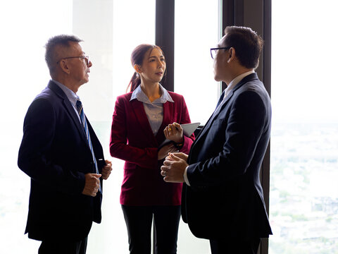 Three businesspeople standing by an office window discussing strategy, meeting, teamwork, leadership, communication, collaboration, planning, management, and professional decisions