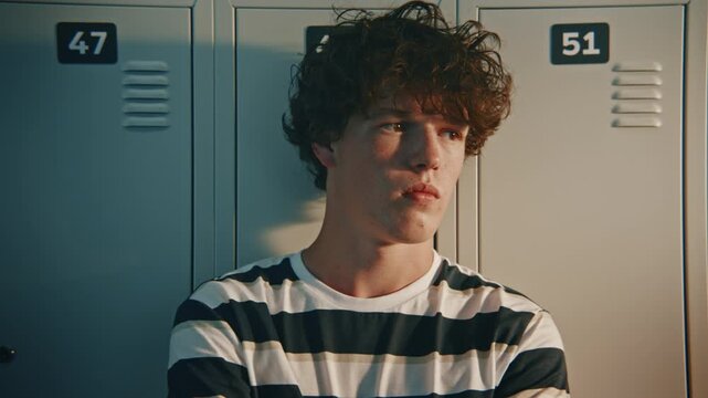 Close Up of Pensive, Curly Haired Teenage Student Staying Alone Near School Lockers Watching Busy Hallway Activity. Concept of Introversion, Teenage Confusion, and Emotional Distance From Classmates.