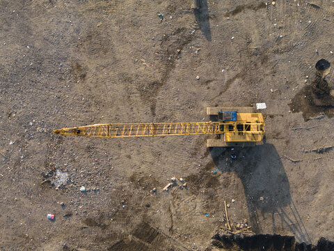 Aerial view of a yellow crawler crane positioned on the dirt ground of the Waste to Energy Facility Construction Project in Kecamatan Denpasar Selatan, Bali, Indonesia.