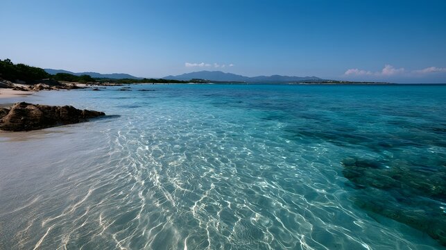 Calm turquoise sea water gently laps over pale sandy shore with distant green hills under clear blue sky